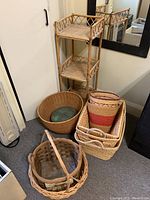 Photo showing three-tier wicker shelving unit standing upright next to several wicker baskets of different shapes, including a large round basket, rectangular baskets with handles, and others stacked.