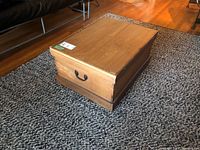 Photo showing rectangular wooden storage box on patterned carpet with metal handle on side and visible wear on top.