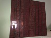 Full frontal view of framed fabric art showing the red textile with detailed woven patterns in vertical stripes of red, green, and black.