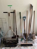 Full view of gardening tools standing upright against a wall, including manual reel lawn mower, shovels, rakes, and brooms.