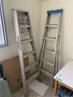 Photo showing two 6-foot folding aluminum ladders standing upright against a wall inside a room.