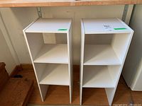 Front view of two white MDF shelving units with three shelves each, placed side by side under a wooden surface.