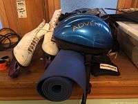 Pair of women's white figure skates size 9 with pink blade guards, blue Advent helmet, and blue rolled yoga mat with carrying strap on wooden surface.