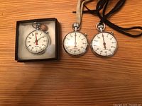 Three silver Minerva stopwatches on wooden surface; one boxed, two with fabric lanyards, one missing glass face.