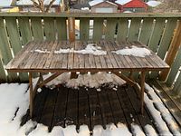 Front view of rectangular wooden outdoor table with slatted top, showing weathering, wear, and minor damage with some snow patches on top and underneath on the deck floor.