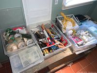 Wide view of three clear plastic bins containing assorted tools such as hand tools, extension cords, pliers, light bulbs, and miscellaneous hardware items.