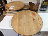 Large round wooden cutting board and other wooden trays stacked on a white surface with plastic tray underneath.