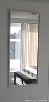 Front-facing photo of a large white framed rectangular mirror mounted on a wall, showing its reflective surface displaying part of a room with vertical blinds and a bench.