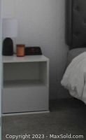 Photo shows one white nightstand with a pull-out drawer and open storage compartment below it, positioned next to a bed with bedding and nearby a lamp and clock on top.
