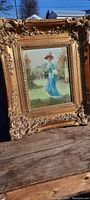 Frontal view of first gilded framed painting depicting lady in blue dress in garden