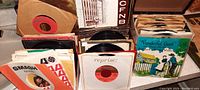 Front view of two small record cases filled with 45 RPM records and one box of additional records, showing colorful and vintage sleeves with various music genres and artists, some sleeved and some not.