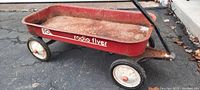 Side profile of the red metal Radio Flyer wagon, showing surface rust on the inside and outside, condition of wheels, and decals on the side.