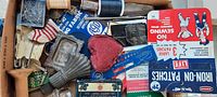 A dresser drawer with assorted vintage sewing items, tinware containers, thread spools, iron-on patches, and a heart-shaped pincushion.