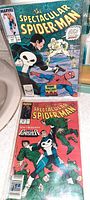 Photo shows two vintage comic books from The Spectacular Spider-Man series with Spider-Man and The Punisher figures on the cover. One comic is priced at 60 cents with Marvel logo and issue details.
