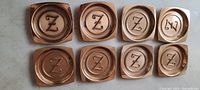 Set of eight antique brass coasters on a surface, showing different shapes with raised letters Z mostly, and one coaster with letter N.