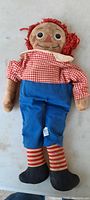 Full front view of vintage Raggedy Ann doll showing facial features, red yarn hair, checkered blouse, blue pants, striped socks, and shoes.