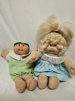Two Cabbage Patch Kids dolls seated next to each other on a white background, showing clothing and general condition.