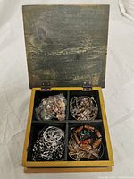 Wooden box opened showing four compartments each containing a collection of assorted costume jewelry including chains, bracelets, and beaded pieces.