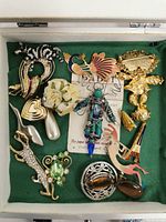 Top view of assorted brooches arranged on green fabric inside the pink display case, showing detailed designs and colors of each brooch.