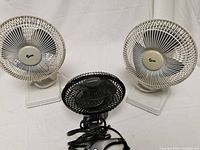 Three small fans displayed on a white surface: two white fans with rectangular bases and one black fan with a rounded base and power cord coiled in front.