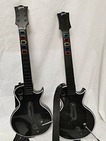 Front of two black Guitar Hero controller guitars, modeled on Gibson Les Paul, showing colored fret buttons and Fender straps attached