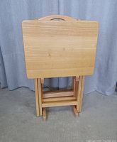 Five wooden folding TV tables stacked and folded for compact storage. Light wood finish with a handle cut-out on tops for easy transport.