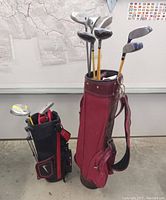 Photo showing two golf bags, a large vintage red golf bag with several long-shafted golf clubs, and a smaller black and red Nike kids bag containing driver and putter clubs.