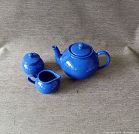Blue ceramic tea set including teapot, sugar bowl with lid, and creamer on a fabric surface.