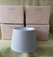Photo of four boxed lampshades with one unboxed gray fabric shade placed in front showing shape and fabric texture.