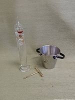 Photo of a Galileo thermometer, stainless steel ice bucket with black handles, and a brass corkscrew on a beige surface.