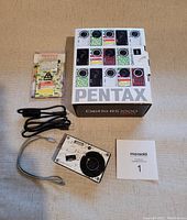 Top view of the Pentax Optio RS1000 camera, USB cable, original box, and packaging material.