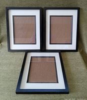 Front view of three black wood picture frames each containing glass and white matting with an inner wooden trim, arranged on a fabric surface.