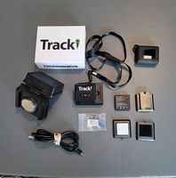 All components of the Tracki GPS Mini Tracker set arranged on a gray surface showing the tracker, magnetic box, batteries, USB cable, and other accessories.