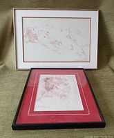 Both charcoal study artworks, one reclining nude with beige matting and silver frame, and a portrait study with red and white double matting in a black frame, placed on green background.