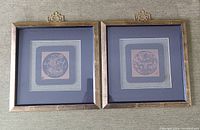 Two framed oriental wood embossed prints side by side on carpet, showing blue double matting and wooden square frames with oriental detail on top.