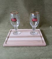 Photo of two clear Stella Artois branded beer glasses with gold rims set on a rectangular wood cutting board with light and medium wood tones.
