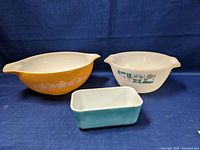 Three Pyrex dishes on blue cloth background: large yellow mixing bowl with white flowers, white casserole with teal/black pattern, small blue loaf dish