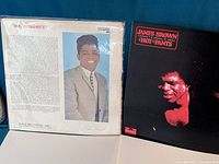Two James Brown vinyl records, 'Hot Pants' and 'Mr. Dynamite'. 'Hot Pants' album on the right has a black cover with a red image of James Brown's face. 'Mr. Dynamite' album on the left has a portrait of James Brown in a grey suit on the back cover with informative text about his career.