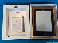 Photo showing Kobo e-reader device in brown protective cover inside original white box with user manual visible under the lid.