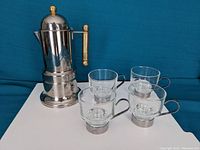 Full view of coffee maker with four demi tasse cups on a white surface and blue background