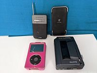 Four electronics items on white surface with blue background: pink iPod with case (cracked screen), silver iPod, black Sanyo cassette player, black AM/FM radio with antenna, and black PowerMat wireless charger