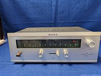 Front view of the Sony FM stereo tuner with analog frequency dial, power switch, tuning knob and control knobs visible on a blue cloth background.
