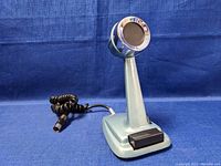 Side angled view showing microphone head with chrome grille, pale blue body and base, coiled cord with connector plug, on a blue fabric background.
