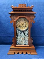 Front view of the wooden vintage pendulum clock showing decorative carved wooden case and floral etched glass panel