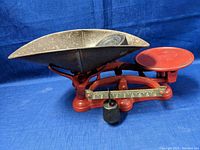 Vintage red base balance scale with metal pans and hanging weight, showing front and side profile.