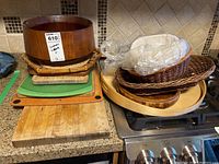 Photo showing assortment of cutting boards stacked on kitchen counter including wooden bowl on top and wicker trays.
