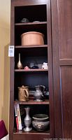 Wooden shelving unit with multiple shelves containing decorated pots, vases, pitchers, figurines, and books.