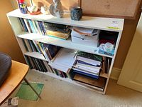 Front view of the white painted wood bookshelf filled with books and office supplies (contents not included).