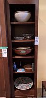 Tall shelving unit with multiple shelves holding various bowls, plates, vases, and decorative items including a large ceramic bowl, wooden bowl, glass bowls, and small vases.
