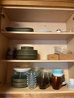 Three-shelf cabinet with stacked Wedgwood stoneware dishes, glass bowls, and ceramic saucers arranged on each shelf.